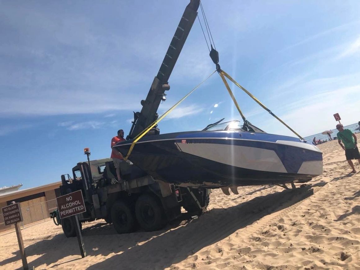 H2O Boat Tow's crane and crew hoisting a boat onto a transport truck at a beach.